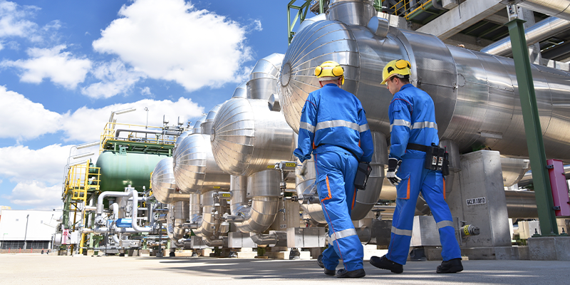 Two industrial workers in blue protective gear inspecting equipment at an energy facility under a bright sky.