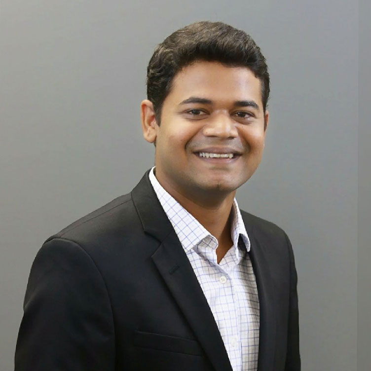 A man in a black blazer and white shirt smiles warmly against a plain gray background, conveying a professional and friendly demeanor.