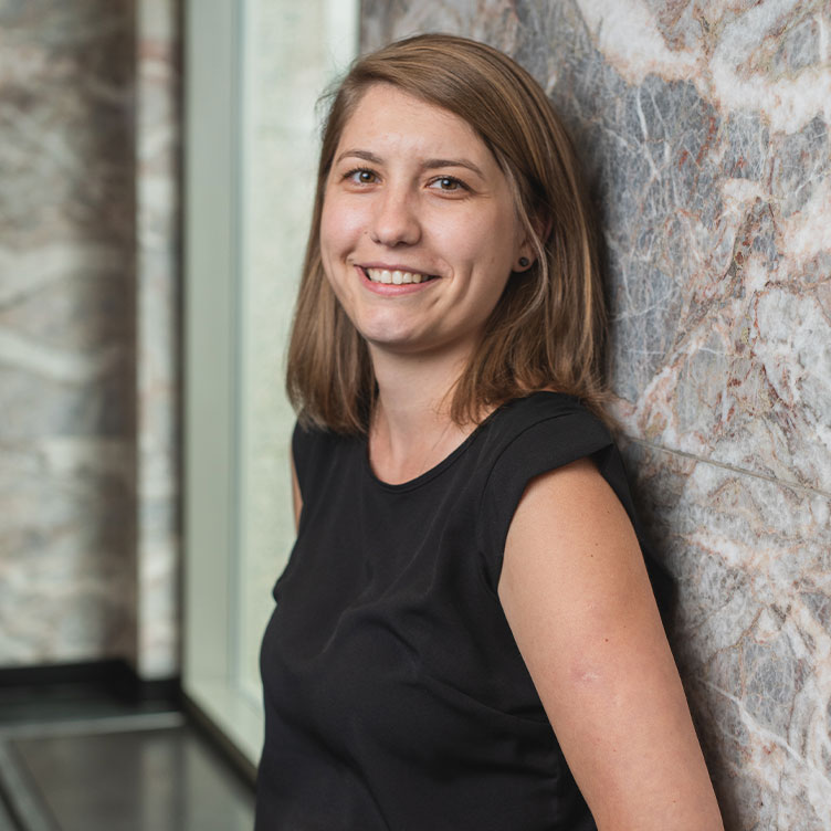 A woman with shoulder-length brown hair and a black sleeveless top smiles, leaning against a textured marble wall, conveying a warm and professional tone.