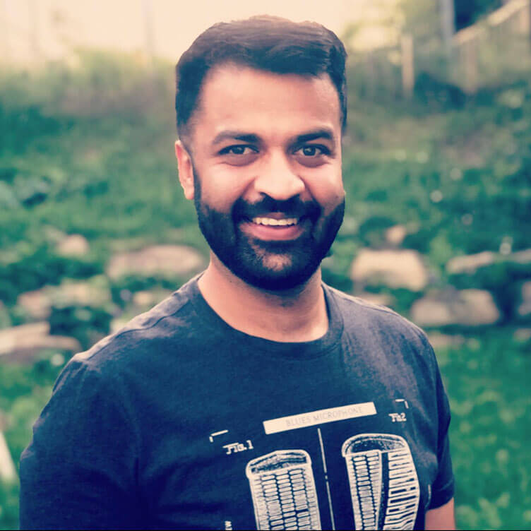 A man with a beard is smiling warmly at the camera, standing in a grassy outdoor setting.