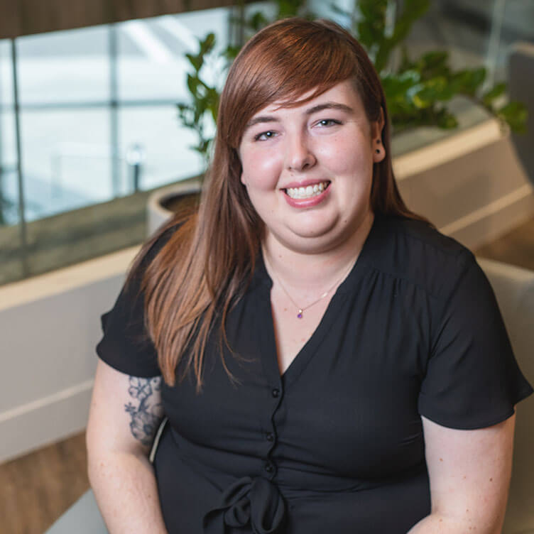 A woman with long, reddish-brown hair sits indoors, smiling warmly. 