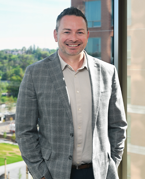 Smiling man in a gray plaid suit stands near a window