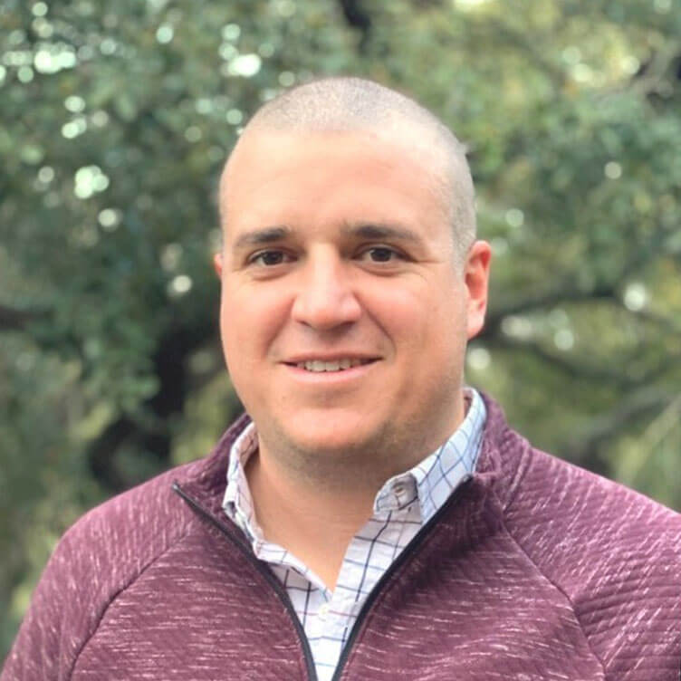 A smiling person in a maroon sweater and checkered shirt stands outdoors with blurred greenery behind them