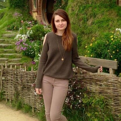 A woman stands by a wooden fence in a lush, green setting, wearing a brown sweater and beige pants. The mood is serene and picturesque, surrounded by flowers.