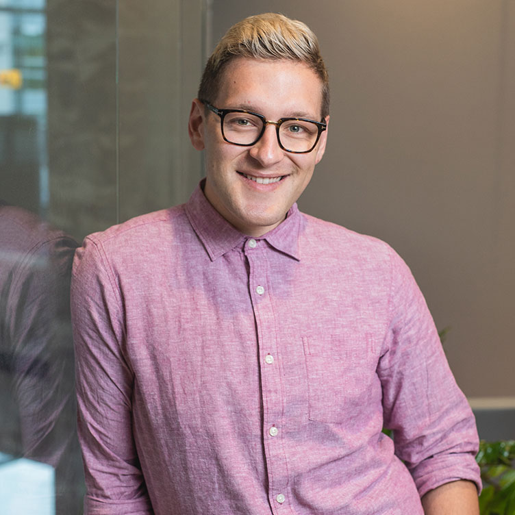 A person with short, light-colored hair and glasses smiles while wearing a pink button-up shirt.