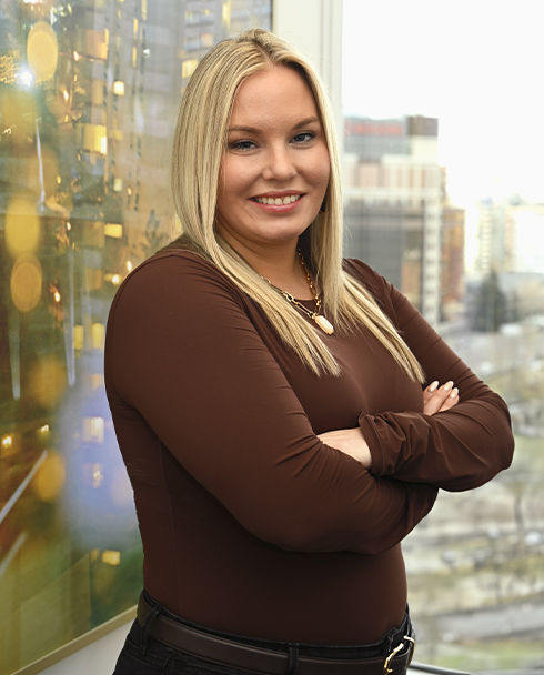 A woman with long blonde hair, wearing a brown top, stands confidently with arms crossed. 