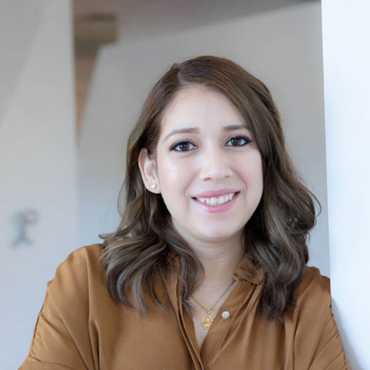 A woman with shoulder-length brown hair smiles warmly, wearing a brown blouse.