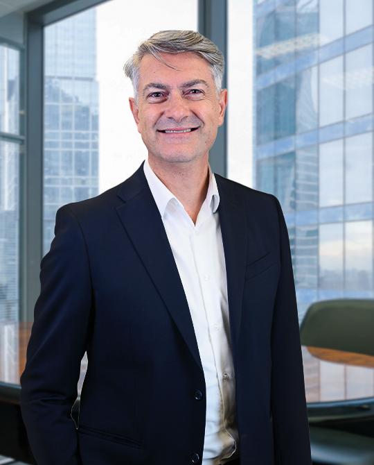 A smiling man in a dark suit and white shirt stands in a modern office with large windows