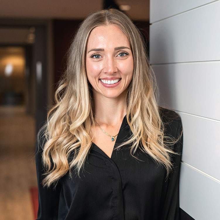 A woman with long blonde hair and a black shirt smiles warmly, standing beside a white wall in a modern hallway with soft lighting.