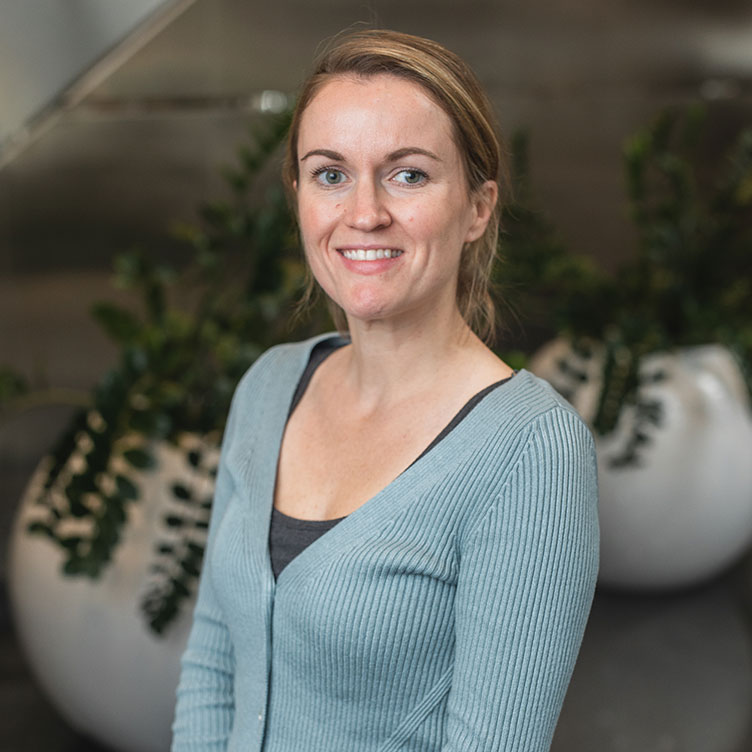 A woman in a light blue sweater smiles at the camera. She stands in front of white planters with greenery, conveying a friendly and welcoming vibe.