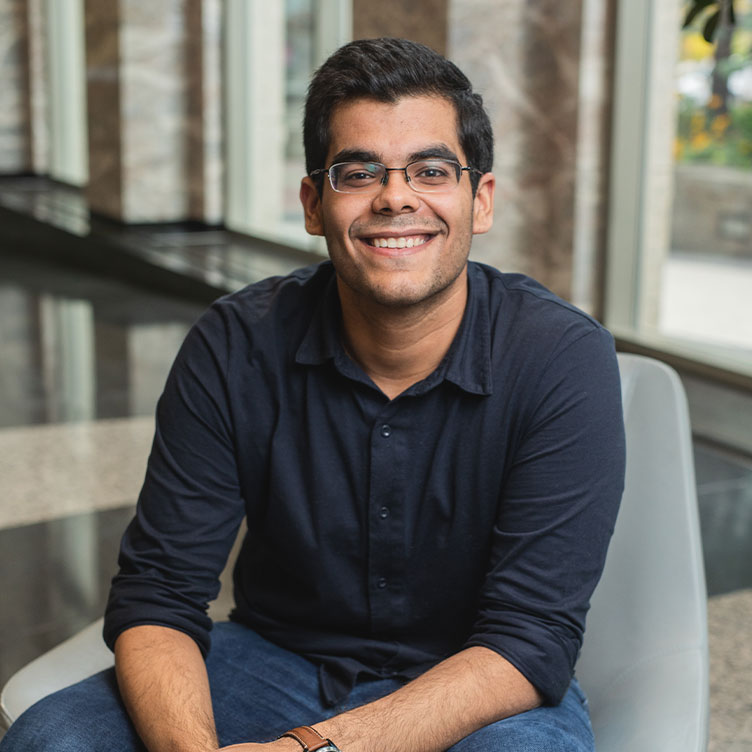 A smiling person in glasses sits casually in a modern setting with large windows and marble floors, wearing a dark shirt and blue jeans