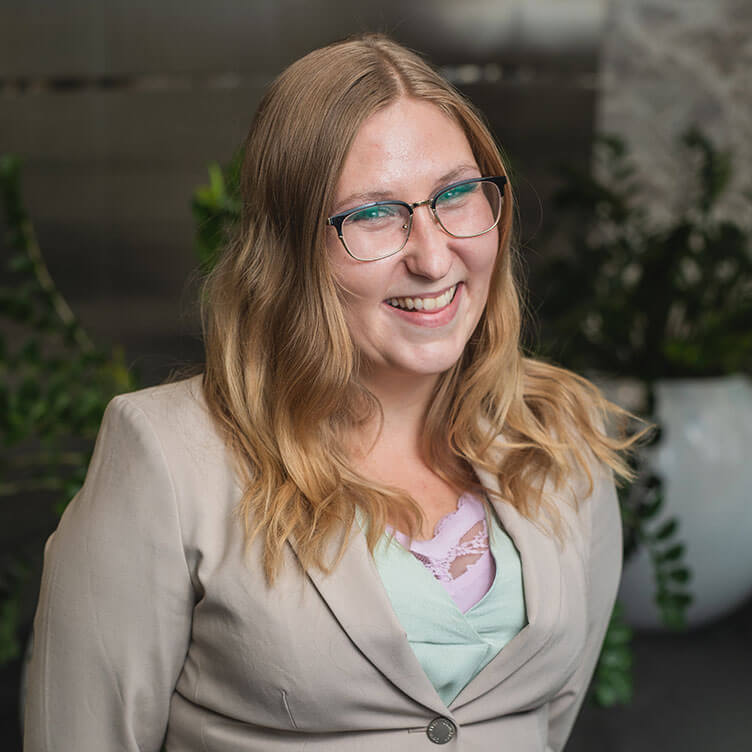 A woman with long blond hair and glasses smiles warmly, wearing a beige blazer over a light green blouse.