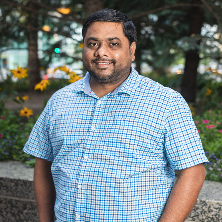 Smiling person in a blue checkered shirt stands outdoors, hands in pockets.