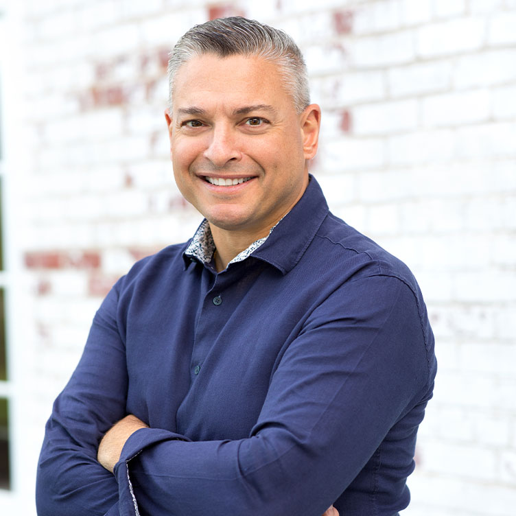 A person in a blue shirt smiles with arms crossed in front of a white brick wall. The tone is friendly and approachable.