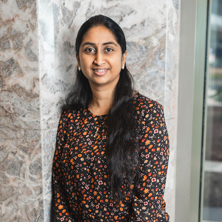 A person with long dark hair smiles warmly, wearing a patterned, long-sleeve top. 