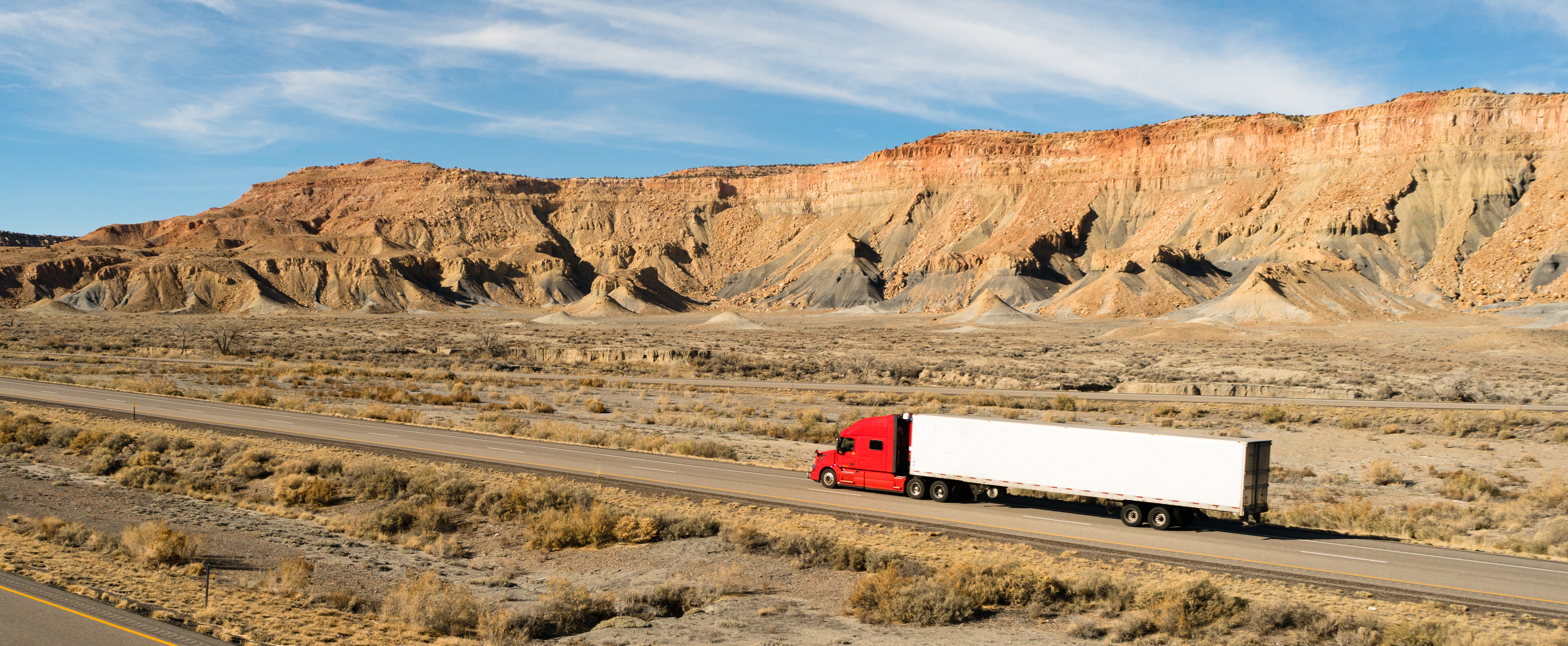 A red semi-truck drives along a barren road, with colorful rocky cliffs rising in the background against a clear blue sky.