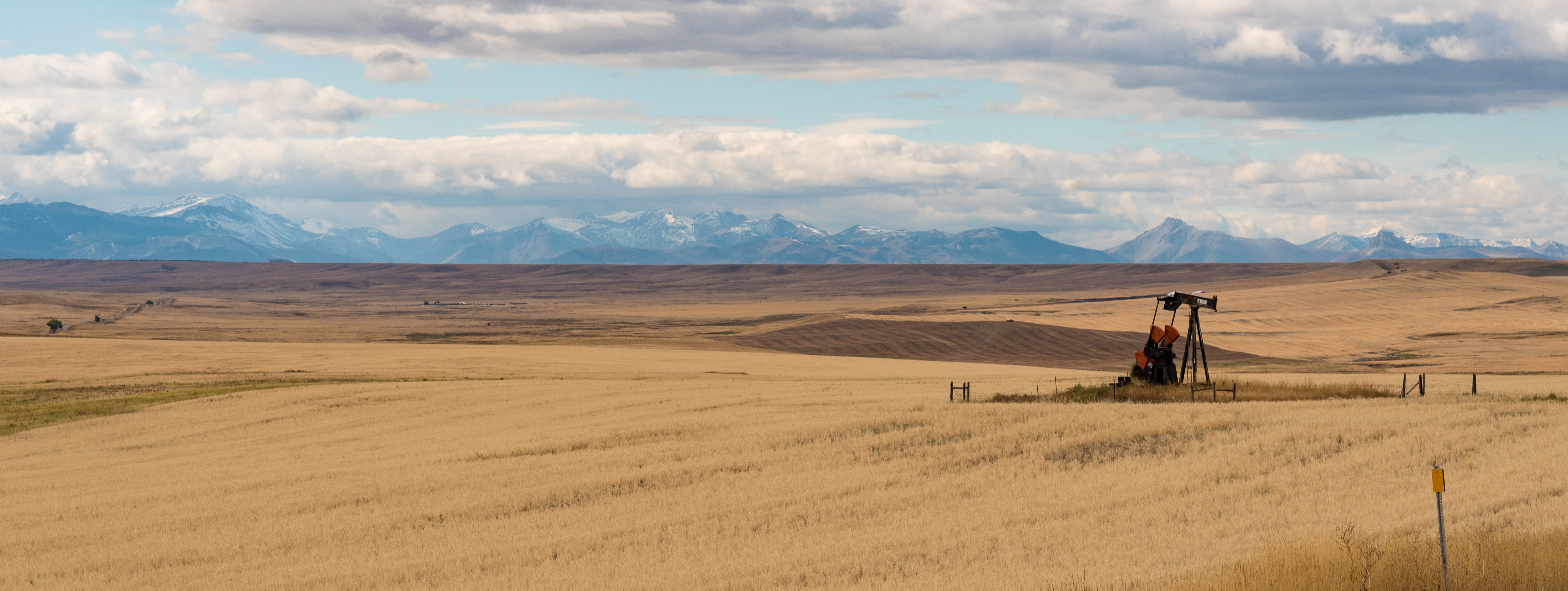 A windmill stands in golden fields under a vast sky, framed by distant mountains and soft clouds on the horizon.
