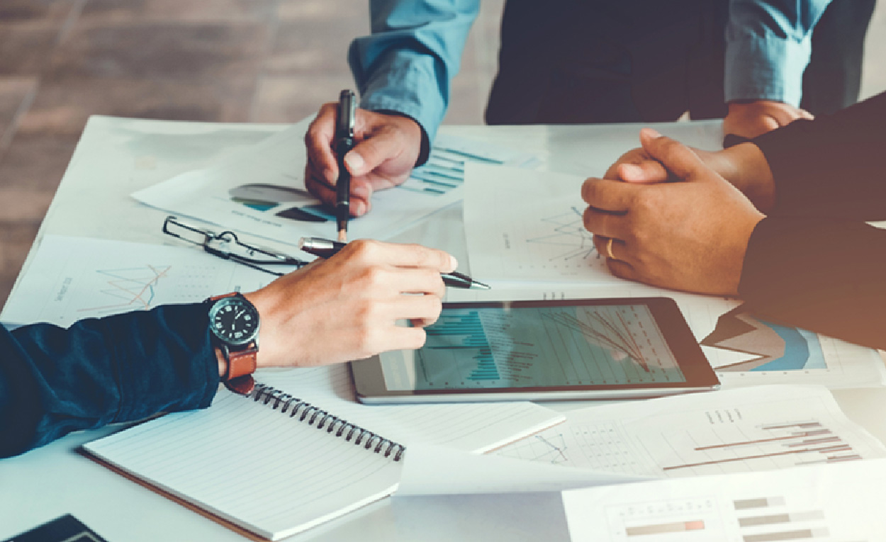 People collaborating at a desk with documents, tablet, and notepad during a business meeting.