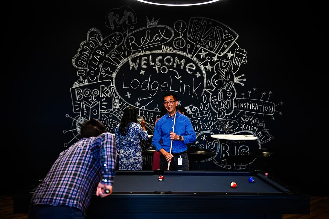 Three people play pool in a dimly lit room. A colorful chalk mural on the wall says "Welcome to LodgeLink," adding a playful, informal vibe.