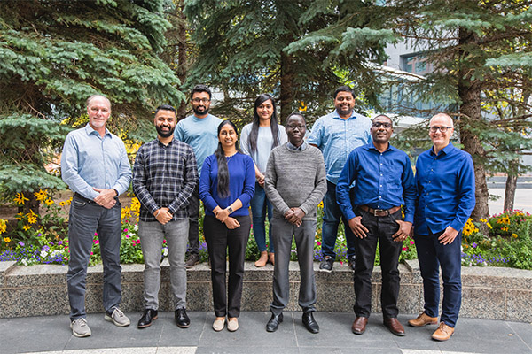 A group of nine people stands outdoors, smiling, with lush green trees and shrubs in the background.