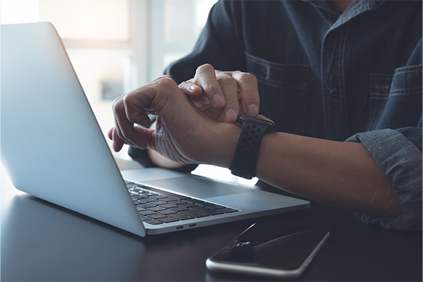 A person in a denim shirt checks a smartwatch while working on a laptop at a desk. A smartphone is placed beside the laptop, suggesting multitasking.