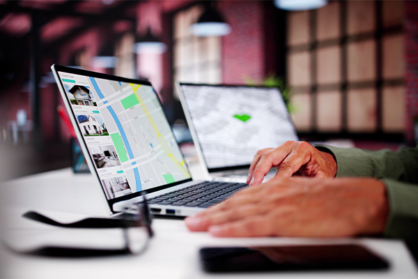 Hands typing on a laptop displaying a map and photos, with another screen in the background. Glasses rest nearby. Modern office setting.