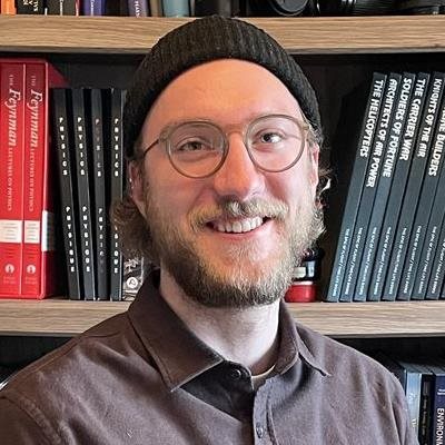 A smiling man wearing glasses and a beanie, standing in front of a bookshelf filled with books.