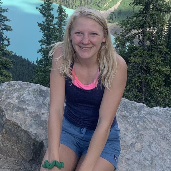 A young woman sitting outdoors on a rock, smiling with a scenic mountain lake in the background.