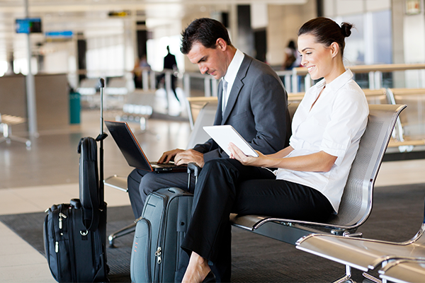 Two business travellers with luggage working on laptops and tablets at an airport.