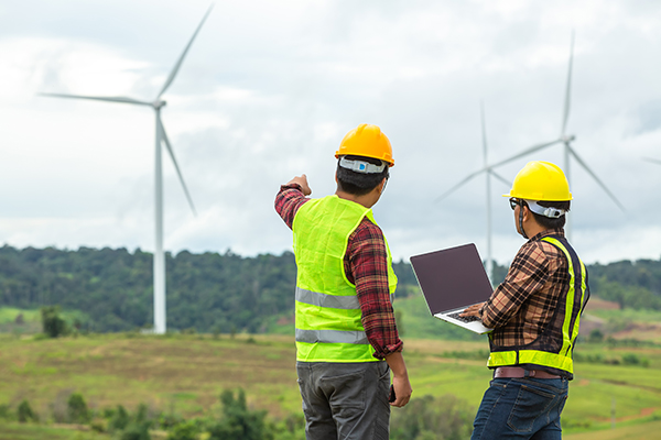two construction workers looking at a laptop in front of wind turbines.	