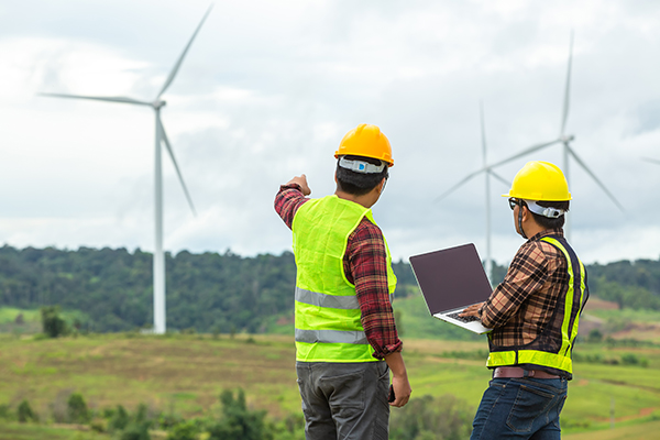 Two construction workers looking at a laptop in front of wind turbines