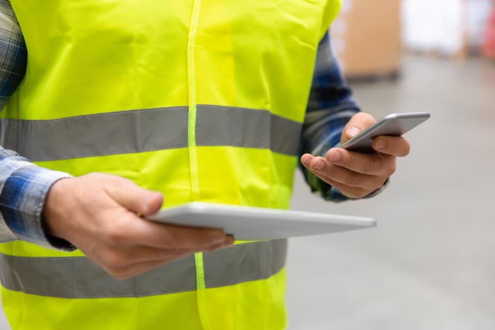 A person in a yellow vest holding a tablet and a phone looking at a workforce travel site